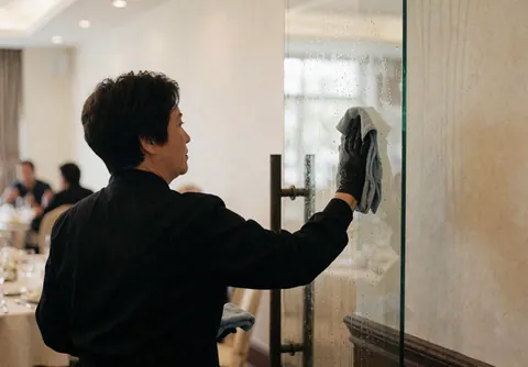 A staff member in uniform cleans glass with a cloth at a banquet. Blounded tables and natural light highlight attention to detail.