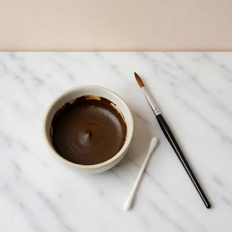 Eyebrow henna in a ceramic container with a brush and cotton swab on white marble for professional application