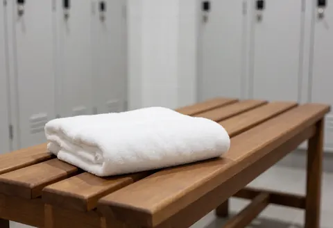 A folded white towel resting on a wooden bench in a locker room with blurred white lockers in the background.