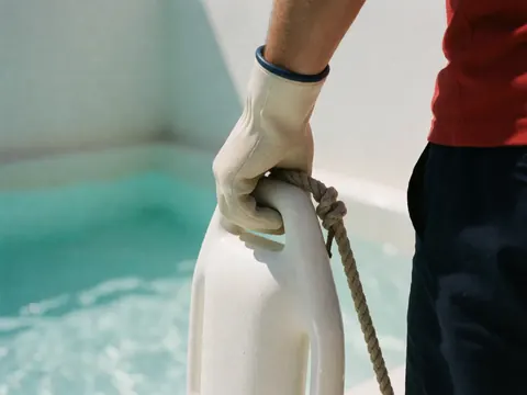 A gloved hand holding a white life ring with a thick rope near a turquoise pool under bright sunlight