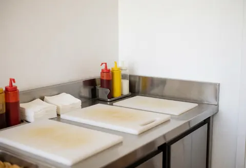 Food preparation station featuring cutting boards with stains, sauce dispensers, and napkins on a stainless steel kitchen surface.