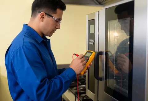 Technician in blue uniform inspects a commercial oven using a multimeter in a kitchen with yellow walls.