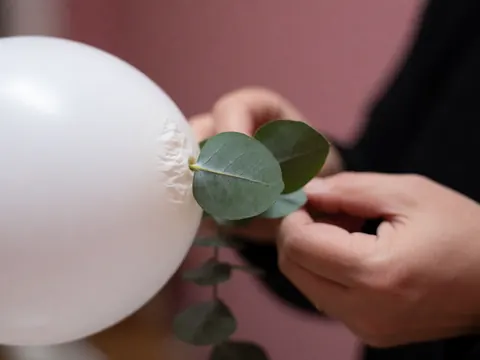 Adult hands placing a green eucalyptus leaf on a white balloon against a soft pink background for event decoration details