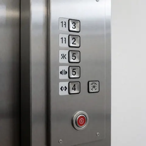Stainless steel elevator control panel with floor buttons and red emergency button mounted on a white wall with soft lighting