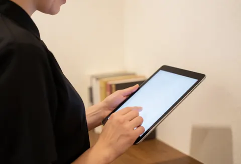 Professional wedding photographer reviewing digital portfolio on a tablet in a warm office setting with blurred books in the background.