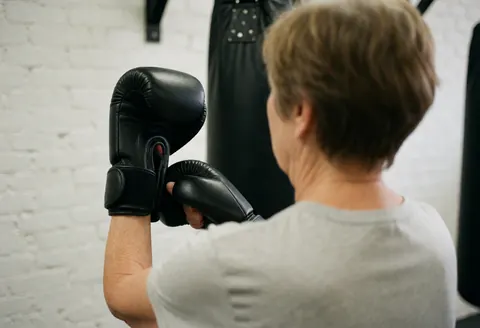 A woman adjusts her black boxing gloves in a gym with a white brick wall and punching bag.
