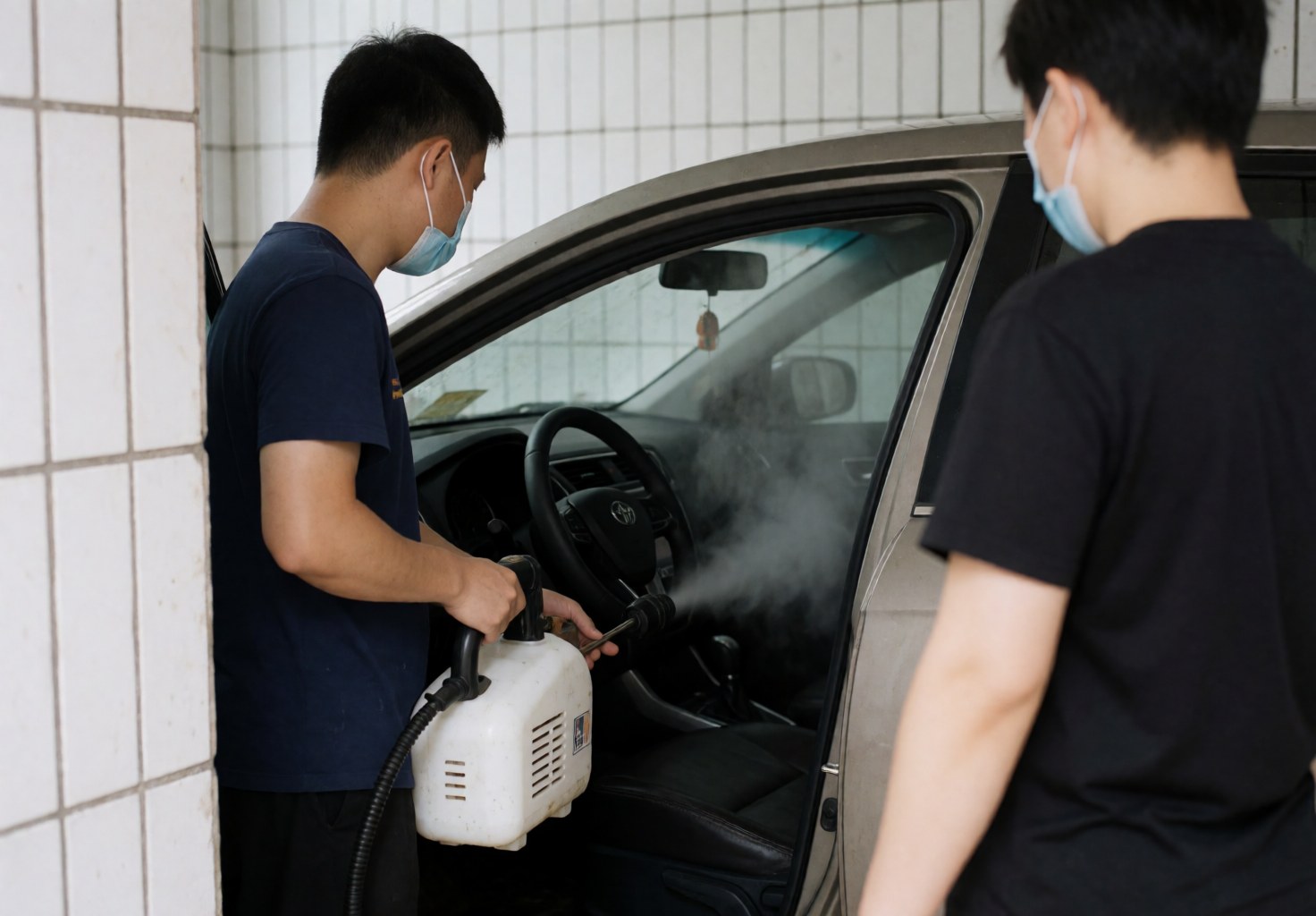 A technician wearing a mask uses a steam machine to disinfect the interior of a car, with the steering wheel and seats visible against white tiled walls.
