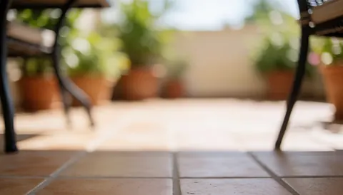 Out of focus patio background featuring a tiled floor, blurred garden furniture, and green potted plants under bright sunlight.