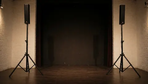 Two professional speakers on tripods stand on a wooden floor against white brick walls in an empty event space with warm lighting.