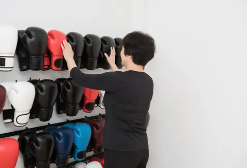 Adult woman in sportswear touching colorful boxing gloves on a shelf in a clean, bright white room.
