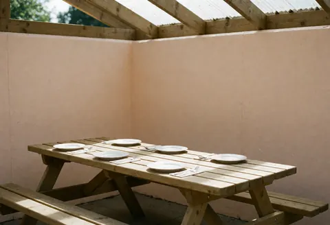 Wooden picnic table under a roof with plates and cutlery ready to eat, beige walls, natural light from the ceiling