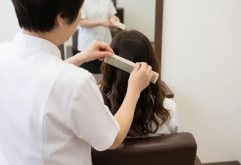A stylist in a white uniform uses a wide-tooth comb to separate curls on a client in a salon with a mirror and brown chair.