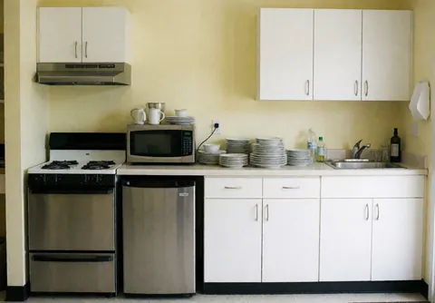Compact apartment kitchen featuring a stove, microwave, refrigerator, and sink with light yellow walls and organized utensils and dishes