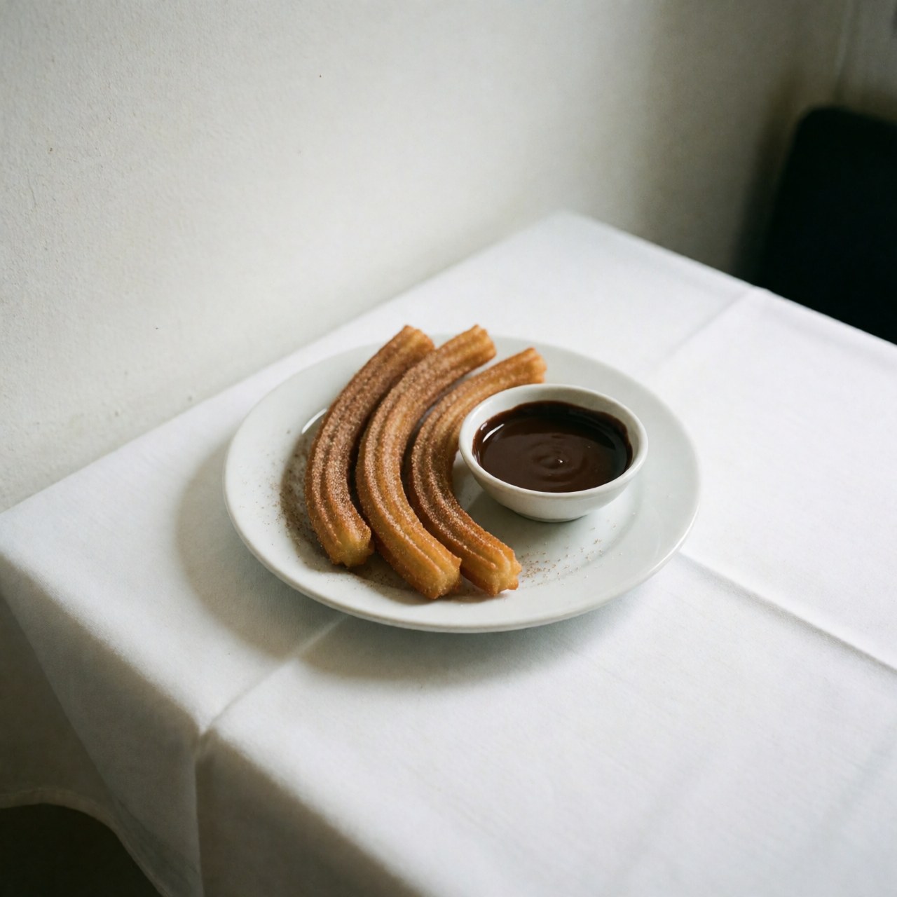 Golden churros dusted with powdered sugar and served with chocolate dipping sauce on a white plate atop a white tablecloth