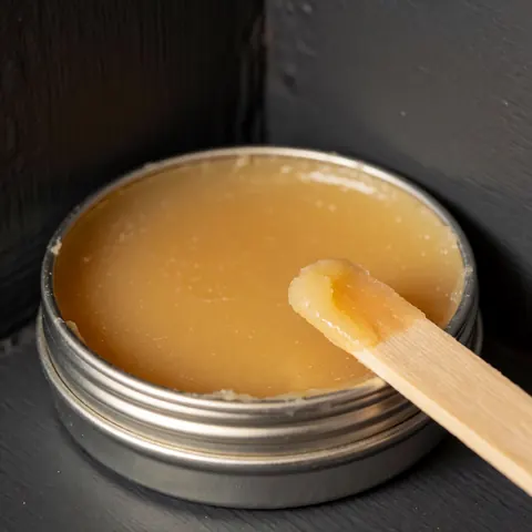 Yellow carnauba wax in a metal tin with a wooden applicator on a gray wall with warm natural lighting