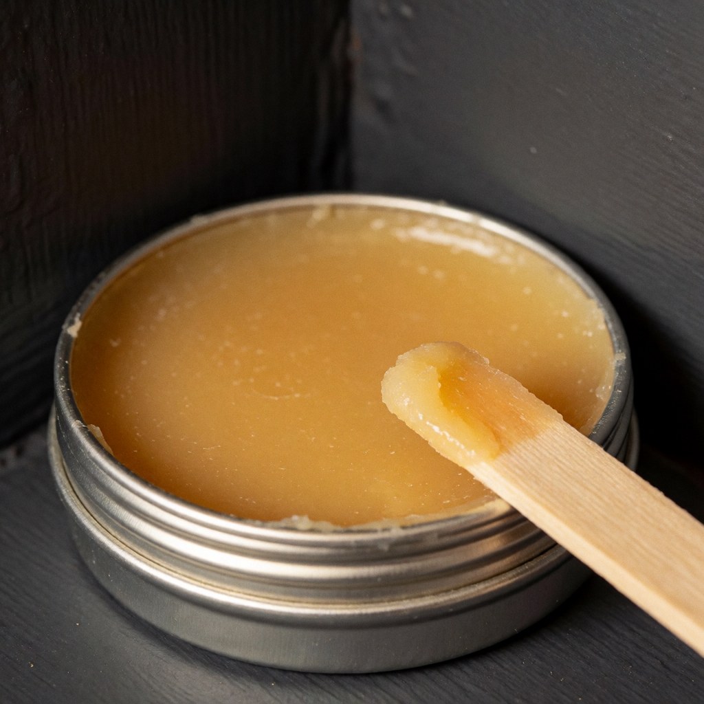 Yellow carnauba wax in a metal tin with a wooden applicator on a gray wall with warm natural lighting