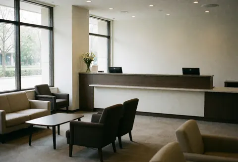 Modern apartment hotel lobby featuring a reception desk with computers, waiting area with sofas and chairs, large windows, white walls, and carpeted floor.