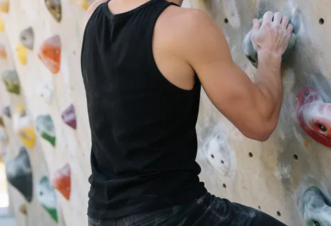 Back view of a climber on a colorful rock climbing wall with chalked hands and defined muscles wearing black athletic gear