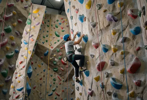 Rock climber in action on a textured artificial wall with colorful holds, visible safety equipment, and gym lighting.