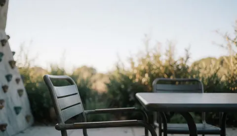 Metal table and chairs on an outdoor terrace with a blurred climbing wall and natural vegetation in the background under soft sunlight.