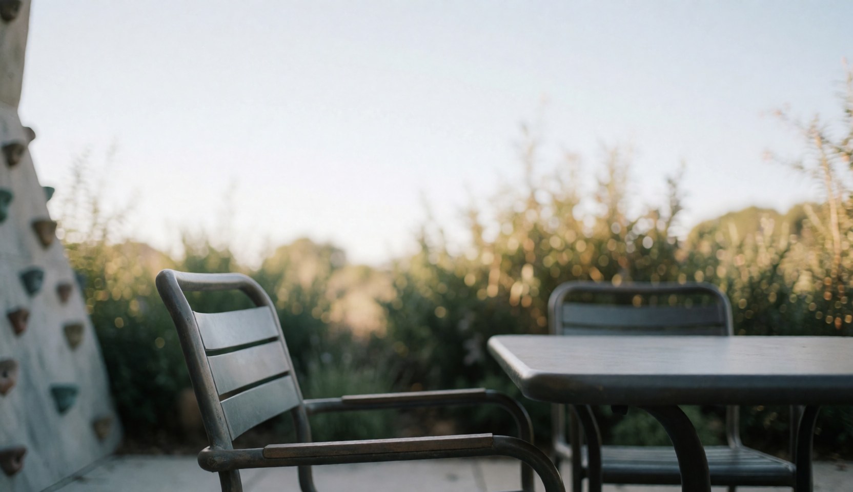 Metal table and chairs on an outdoor terrace with a blurred climbing wall and natural vegetation in the background under soft sunlight.
