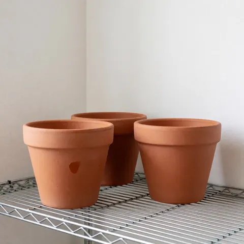Three unpainted terracotta flower pots resting on a metal shelf against white walls with soft morning light highlighting the natural texture