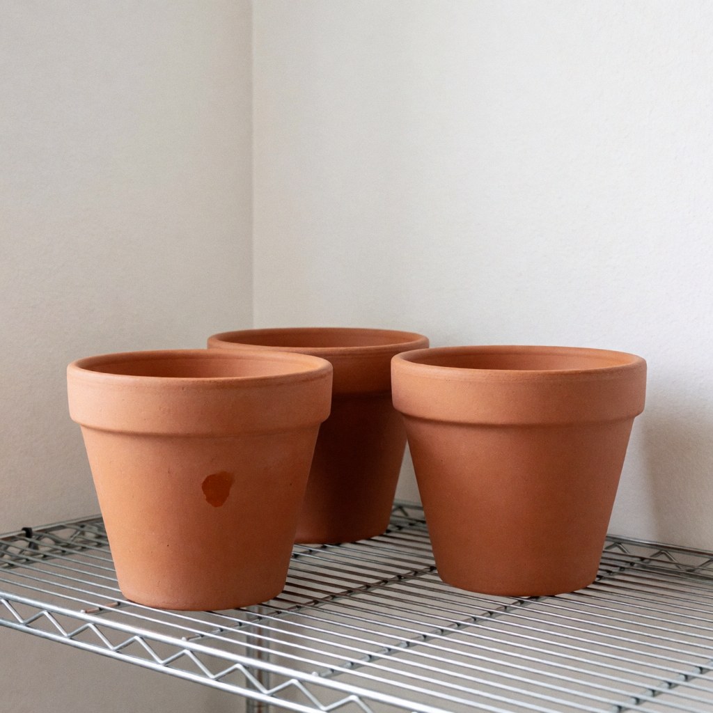Three unpainted terracotta flower pots resting on a metal shelf against white walls with soft morning light highlighting the natural texture