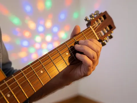 Close-up of a hand strumming a guitar string under festive party lights with colorful bokeh on a white background.