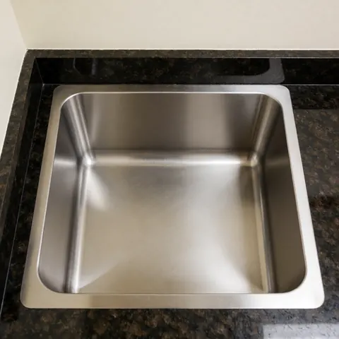 Top view of a brushed stainless steel kitchen sink installed in a dark granite countertop with bright lighting and warm white walls
