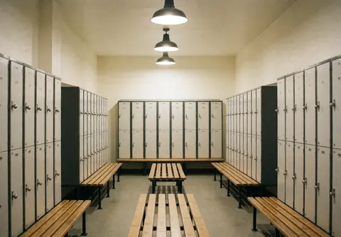 Empty locker room featuring metal lockers and wooden benches illuminated by hanging lamps, ready for visitors.