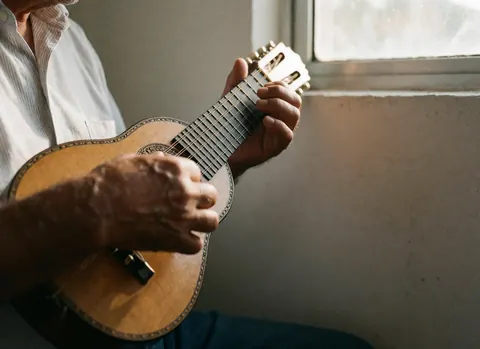 Close-up of hands playing a charango, showing strings and polished wood against a white wall in a warm, professional setting.