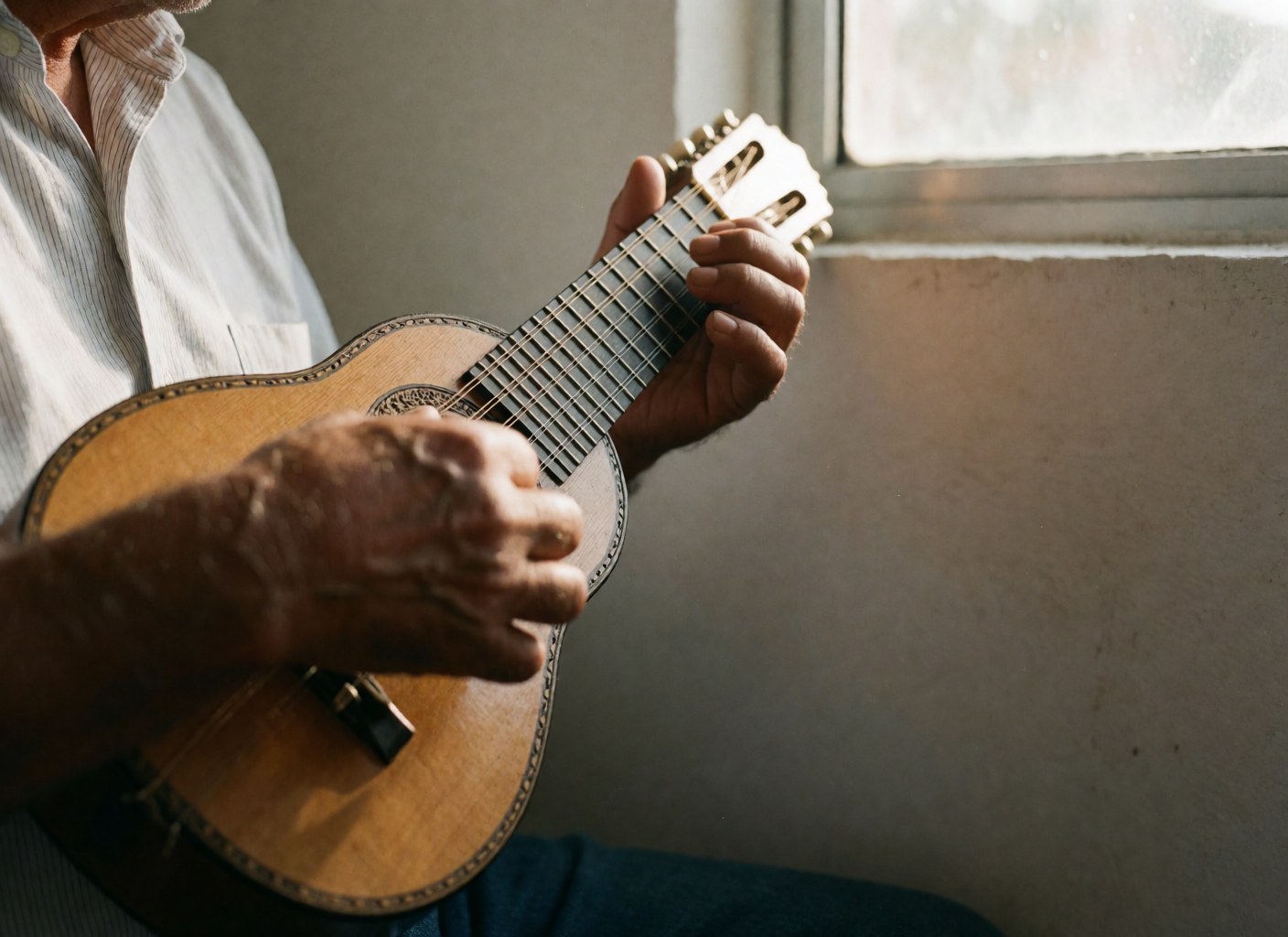 Close-up of hands playing a charango, showing strings and polished wood against a white wall in a warm, professional setting.
