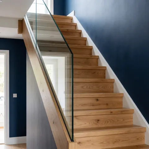 Polished oak staircase featuring a transparent glass railing, set against dark blue walls with natural light in a modern interior hallway.