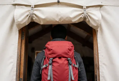 A hiker wearing a red backpack and gray jacket enters a mountain refuge through a wooden door with a beige canvas awning, revealing an interior with wooden beams.