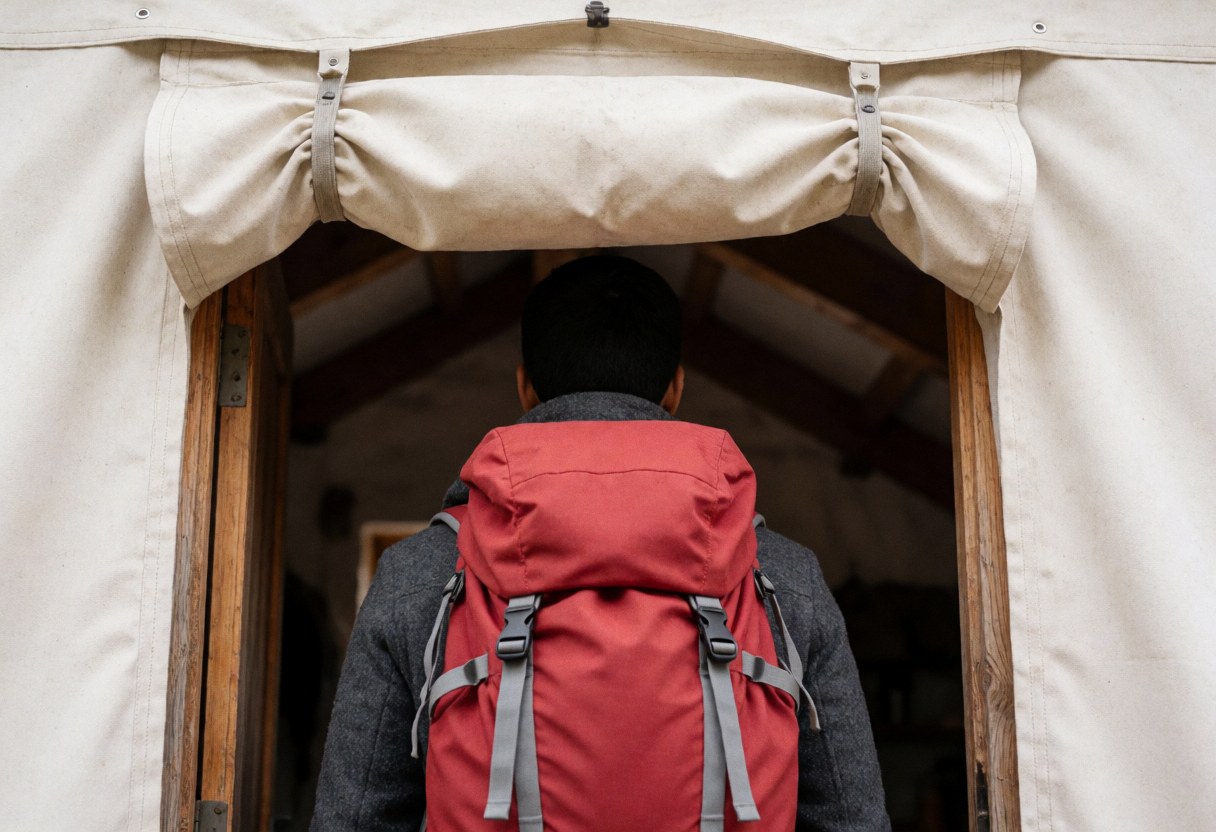 A hiker wearing a red backpack and gray jacket enters a mountain refuge through a wooden door with a beige canvas awning, revealing an interior with wooden beams.