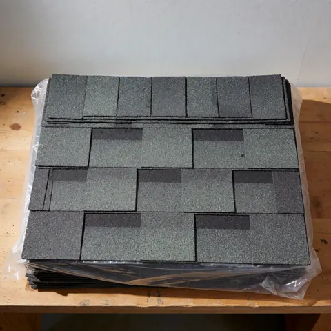 A plastic-wrapped package of textured gray asphalt shingles resting on a wooden bench against a plain wall background