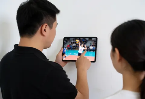 Volleyball coach analyzing attack video on a tablet while another person observes from behind in a room with white walls