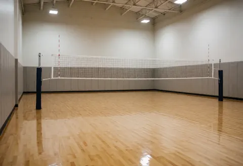 Empty volleyball court with polished wooden floor, white net, black posts, safety wall panels, and ceiling lighting