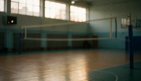 Blurred indoor volleyball court background featuring a net, posts, and wooden floor illuminated by warm light from high windows.