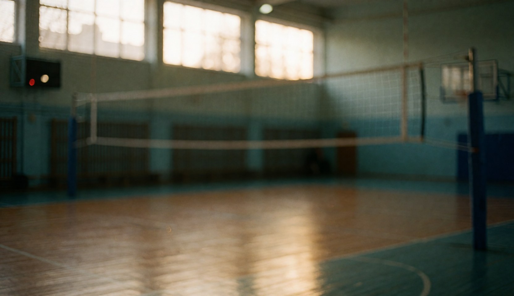 Blurred indoor volleyball court background featuring a net, posts, and wooden floor illuminated by warm light from high windows.