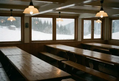 Empty mountain lodge interior featuring rustic wooden tables, benches, hanging lamps, and windows showing falling snow.