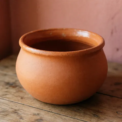 Matte finish artisanal clay cazuela resting on a rustic wooden surface with a pink wall background and natural lighting highlighting the visible clay texture