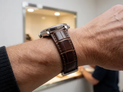Close-up of a watch clasp with a crocodile leather strap against a lit display case background, highlighting leather texture and metallic finish.