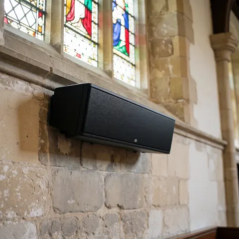 Black linear speaker mounted on stone wall in church with colorful stained glass and natural light filtering through