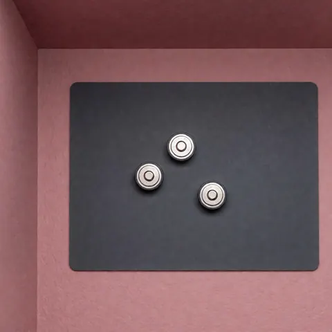 Three silver button cell batteries resting on a gray anti-static pad against a pale pink wall, suitable for watch repair and electronics work