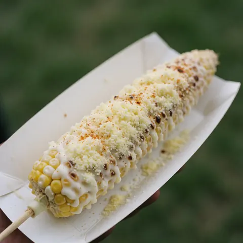 Grilled corn topped with white mayonnaise, shredded cheese, and spices, resting on white paper against a green background.