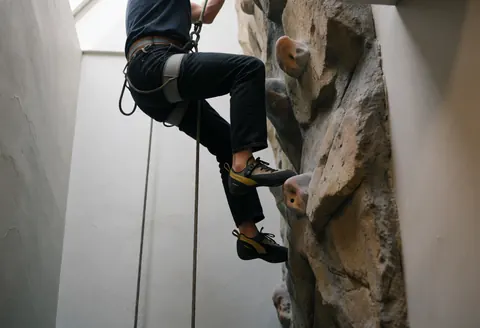 Person scaling an artificial rock wall with climbing gear, ropes, and harness under natural light in an adventure park setting
