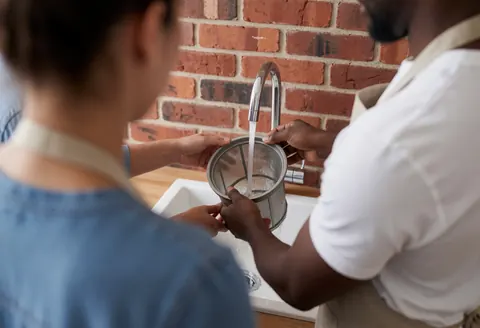 Technician cleaning dishwasher filter under running water in a white sink with a red brick wall background