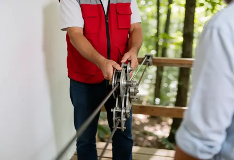 Guide in red vest adjusts cable pulley on wooden platform with blurred forest background and natural light
