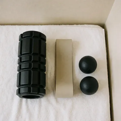 Black foam roller, beige resistance band, and massage balls arranged on a white towel for athletic recovery and training.